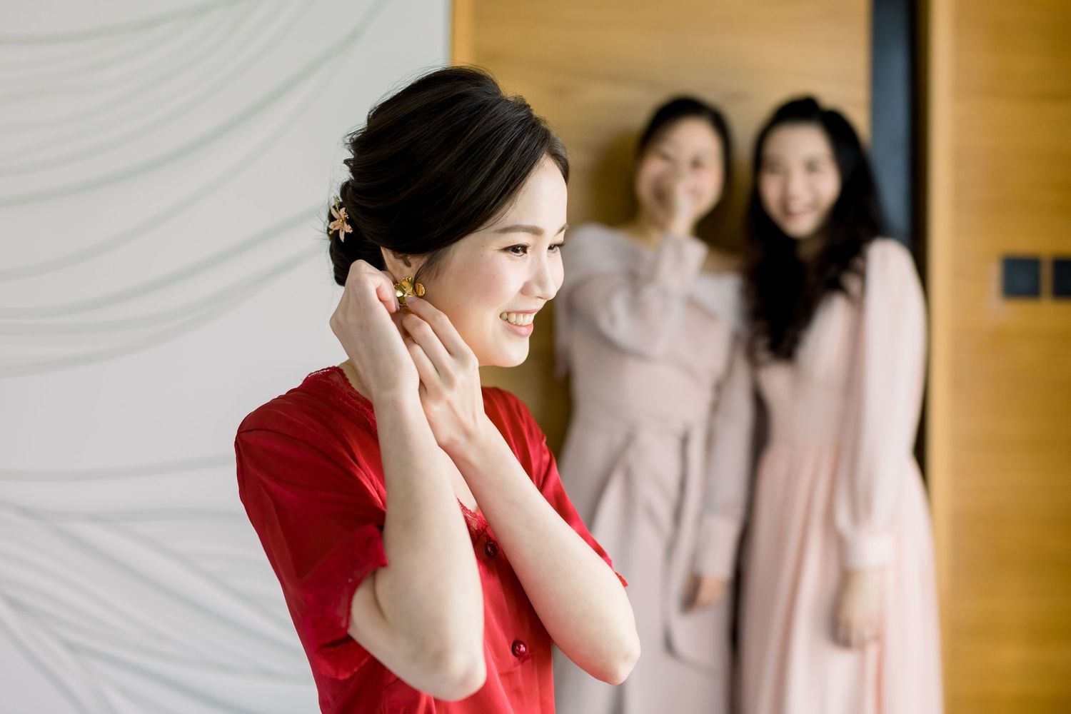 Three people in traditional Asian dresses share an emotional moment against a wooden wall.