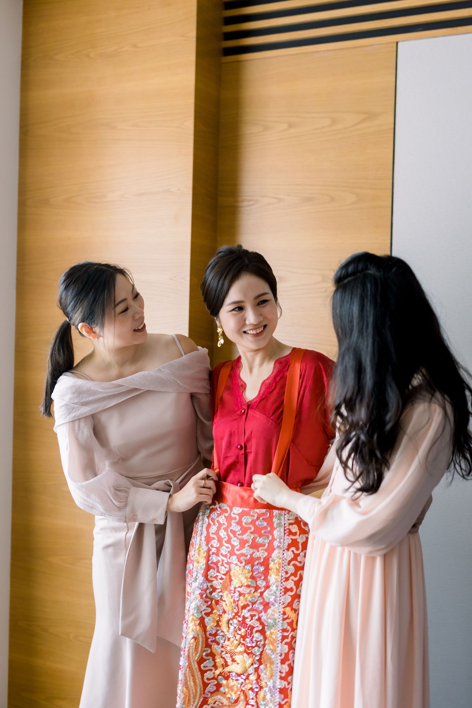 Group wearing traditional Chinese dresses sharing joyful moments against wooden paneled wall.