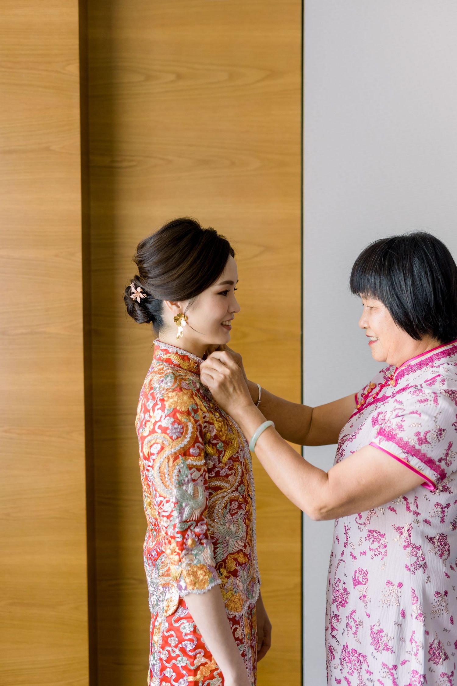 Two people in floral qipao dresses interact in front of wooden panels.