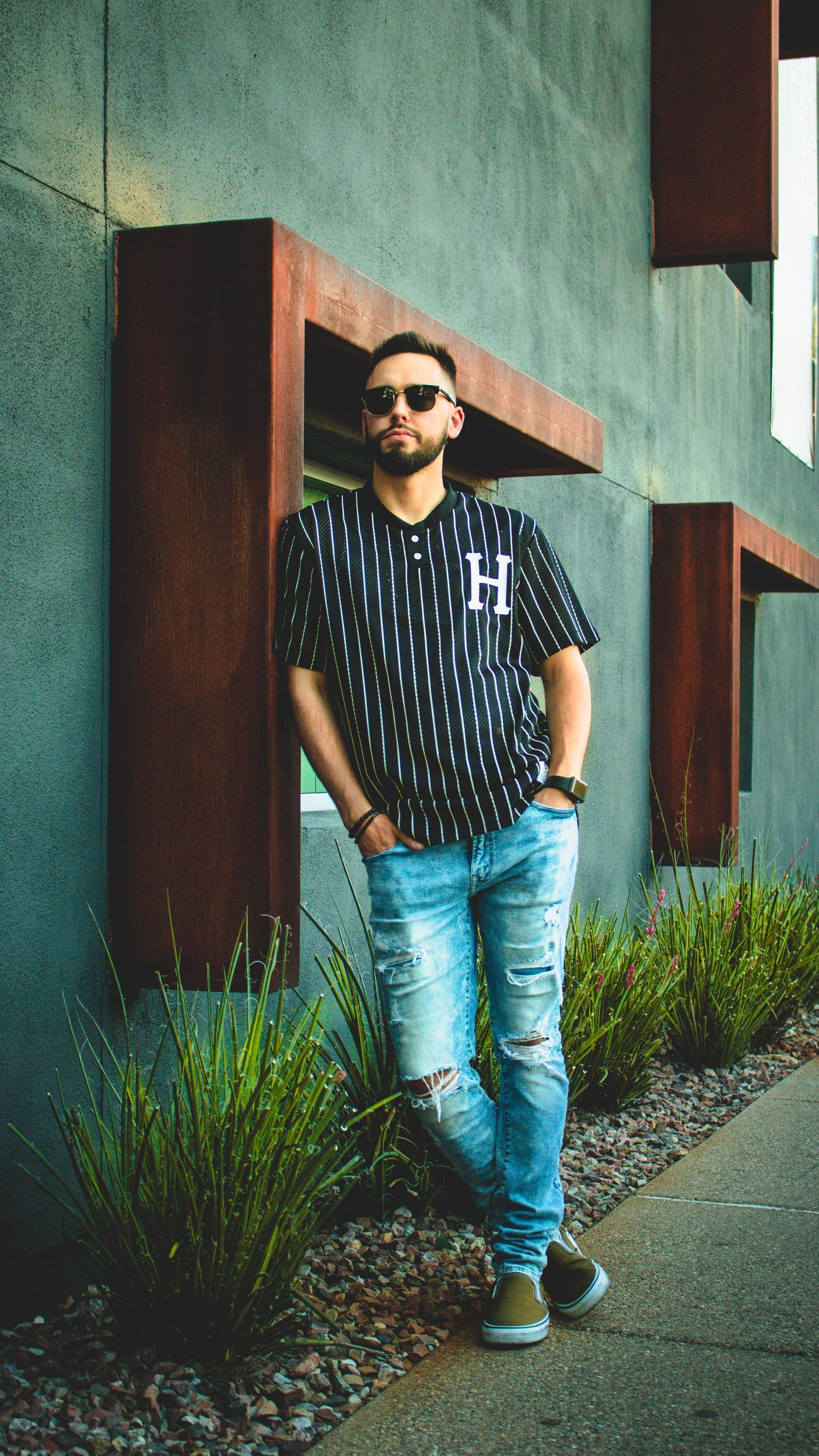 Person in pinstripe baseball jersey and ripped jeans poses against teal wall with architectural details.