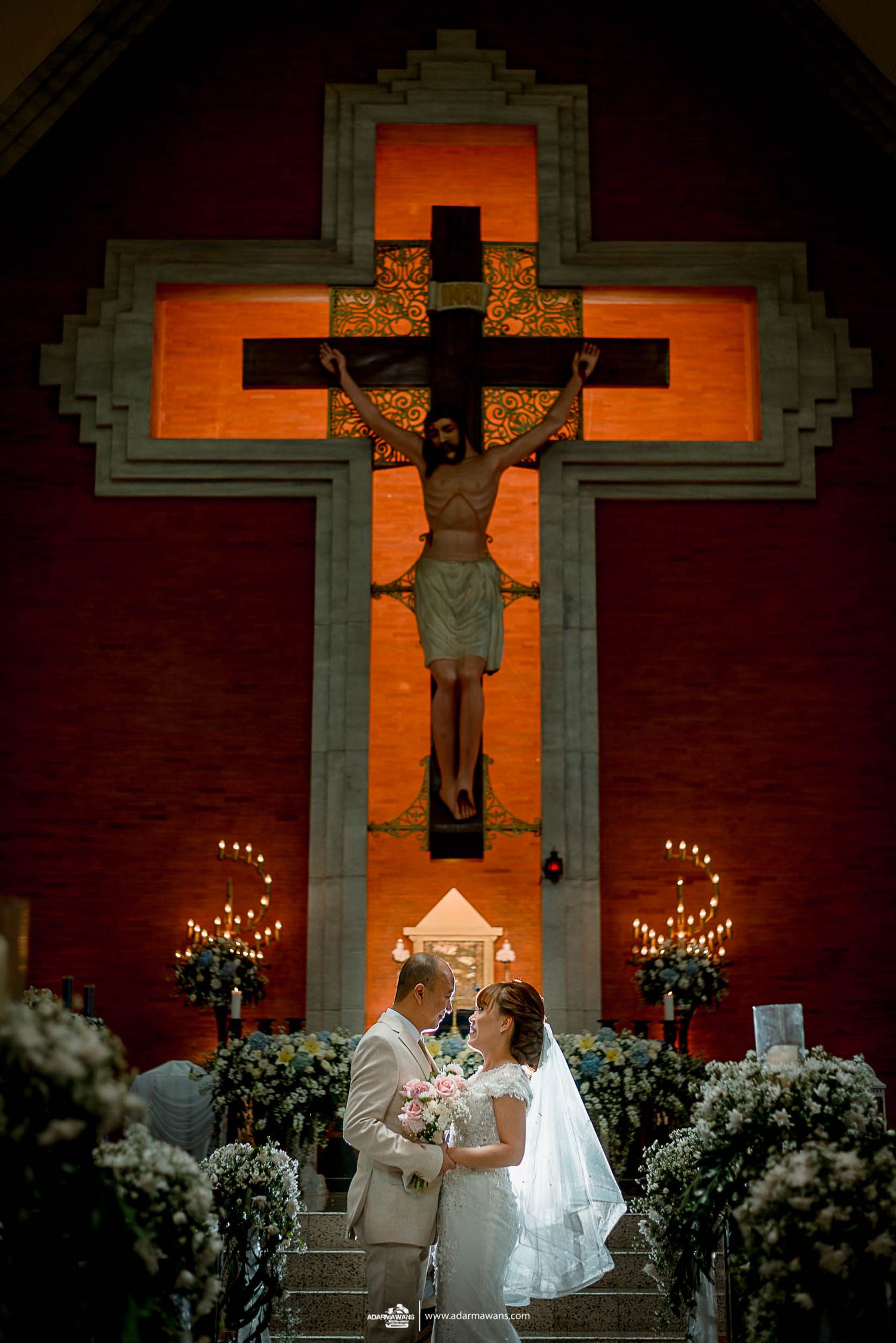 Wedding ceremony in church with dramatic illuminated crucifix and floral decorations.