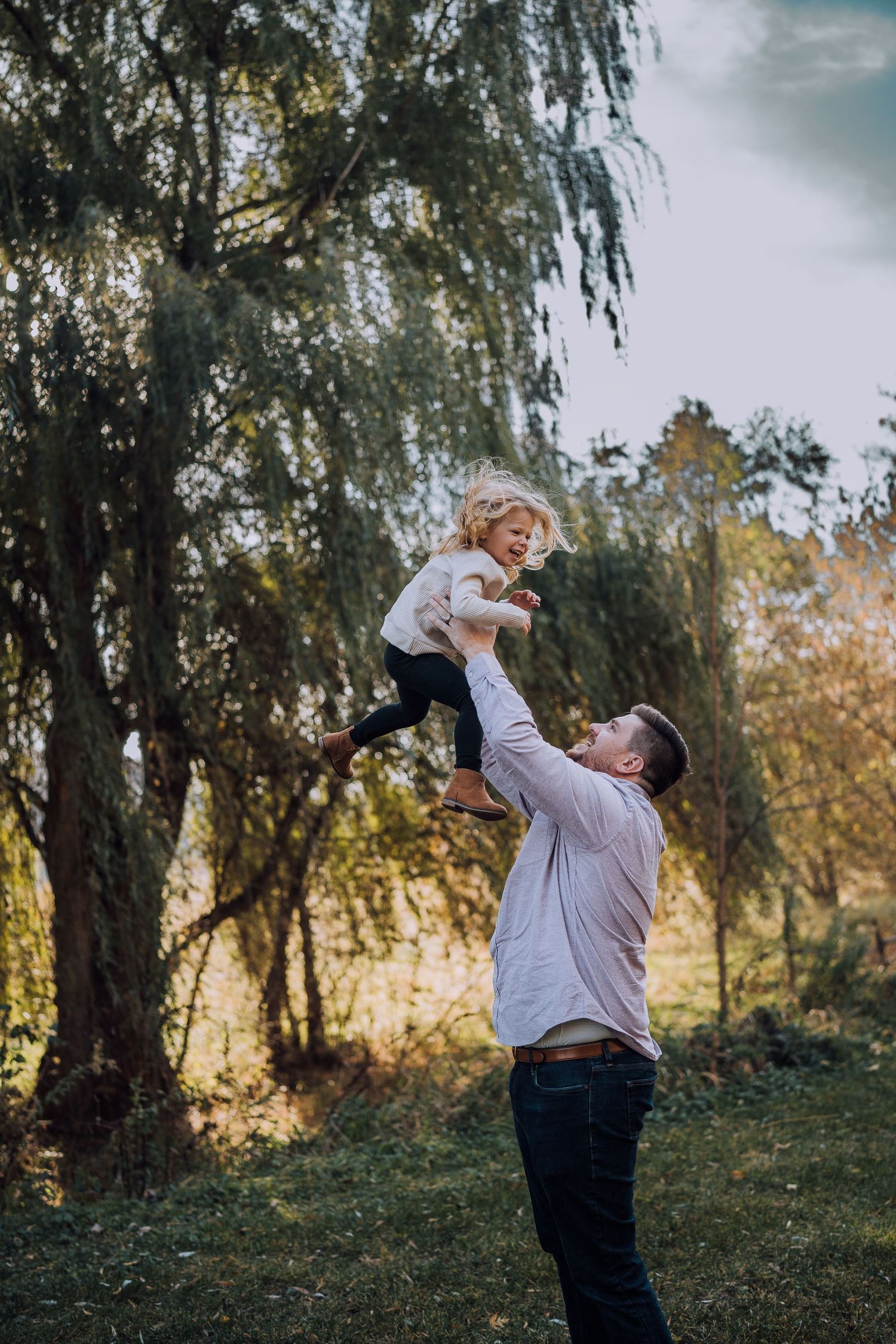 A playful moment captured as a child is lifted high into the air under a willow tree.