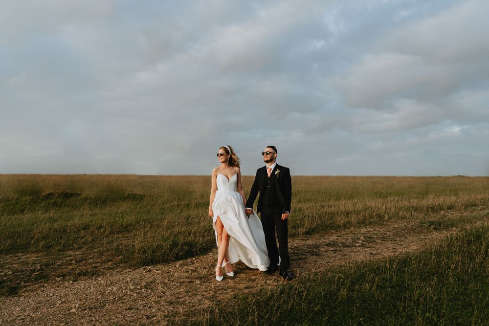 A wedding couple walks hand in hand through an open field under a cloudy sky.