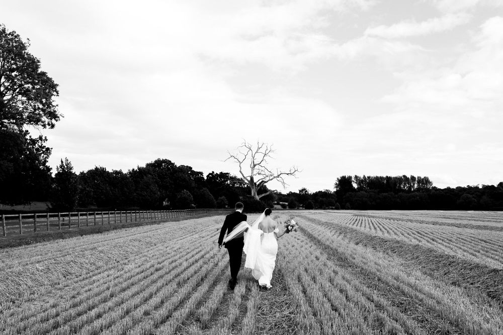 Black and white image of couple running joyfully through a plowed field with fence in background.