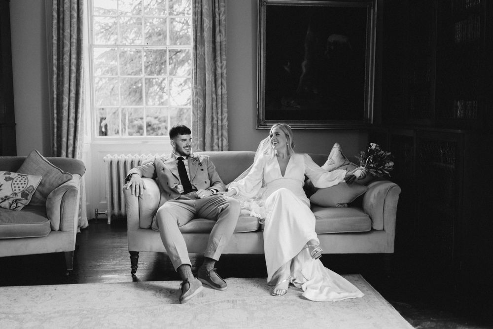 Black and white photo of a relaxed wedding couple seated on elegant sofas indoors.