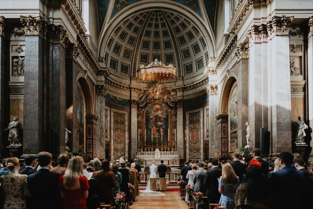 Interior of a grand cathedral during a ceremony with ornate baroque architecture and domed ceiling.