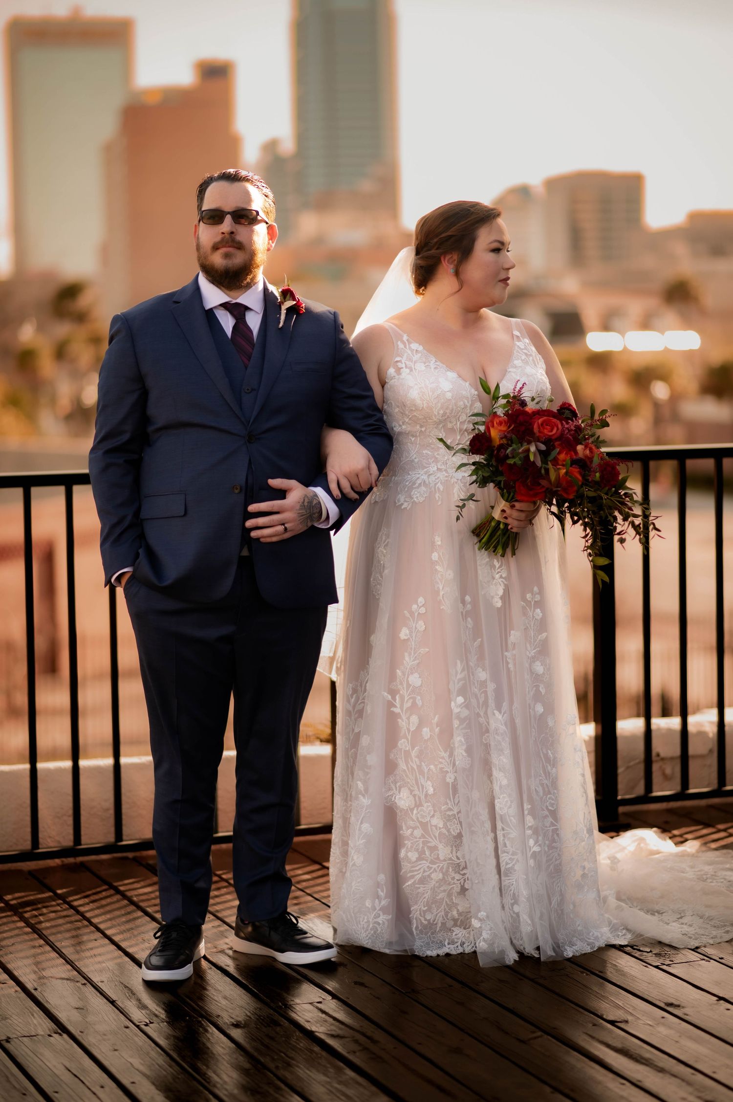 Newlywed couple posing on wooden deck against urban skyline at sunset.