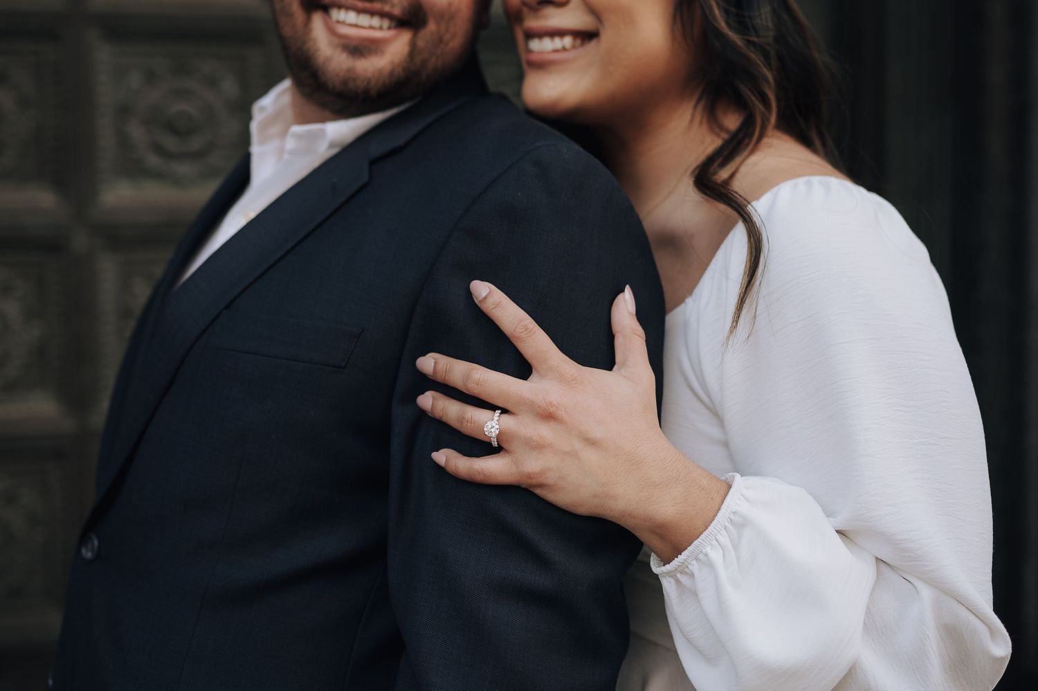 Close-up of couple in formal wedding attire showing engagement ring against dark suit jacket.