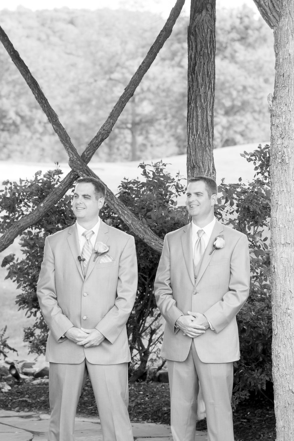 Groomsmen in light gray suits stand under wooden pergola during outdoor ceremony.