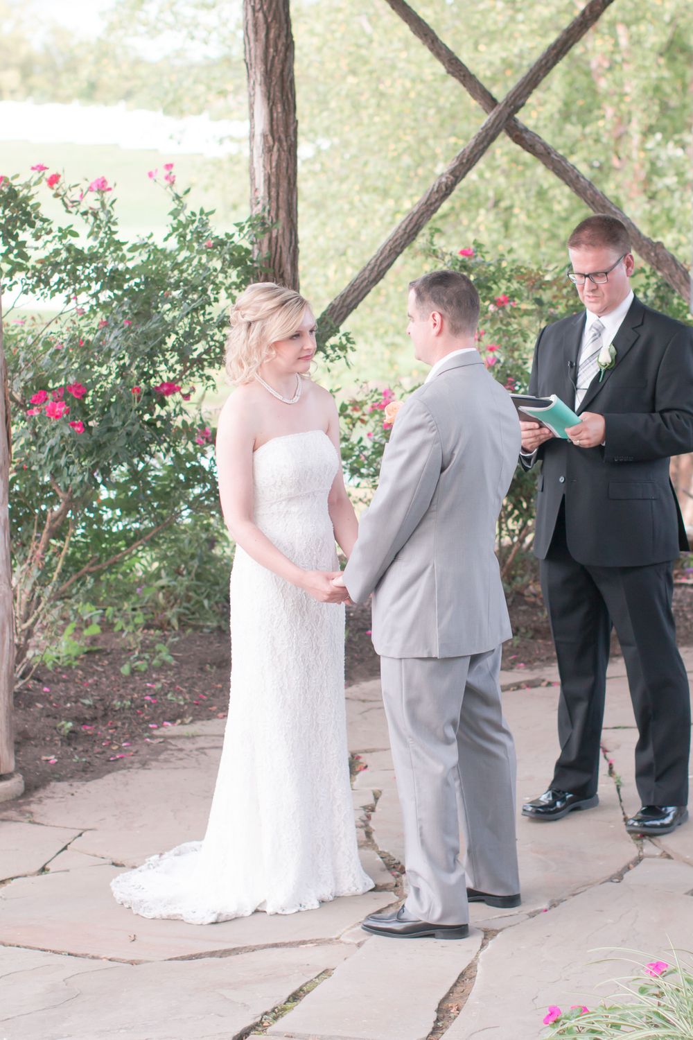Bride and groom stand before officiant during outdoor ceremony under rustic wooden structure.
