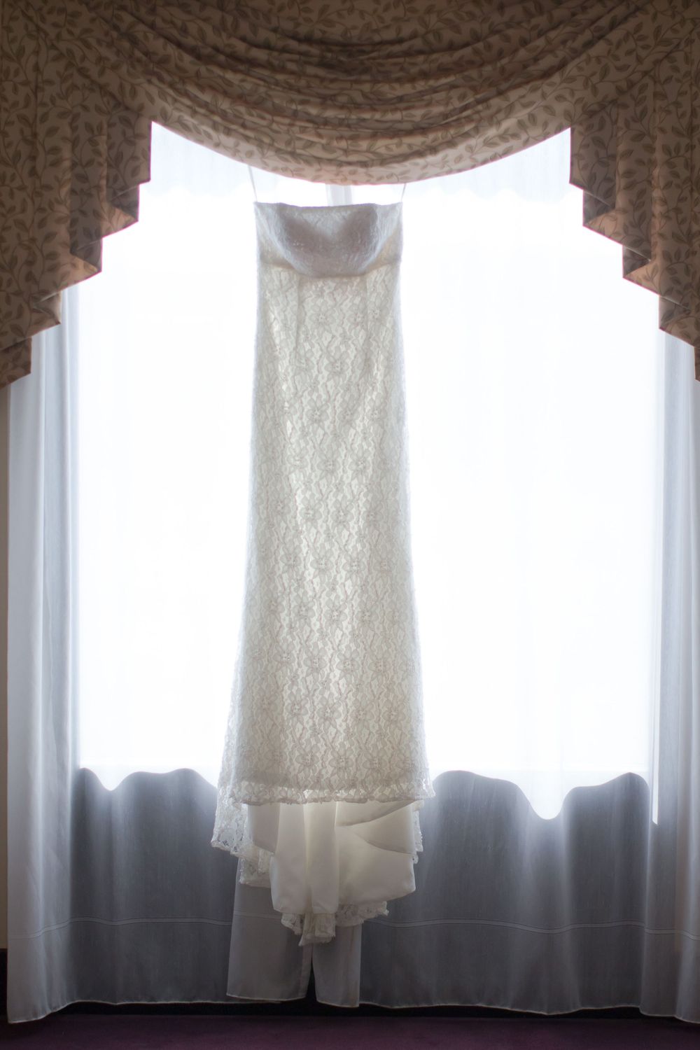 Lace wedding dress hanging in window silhouette with elegant draped curtains.