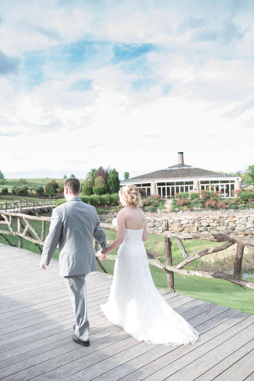 Newlyweds walk along a wooden bridge towards an elegant venue with lush green landscaping.