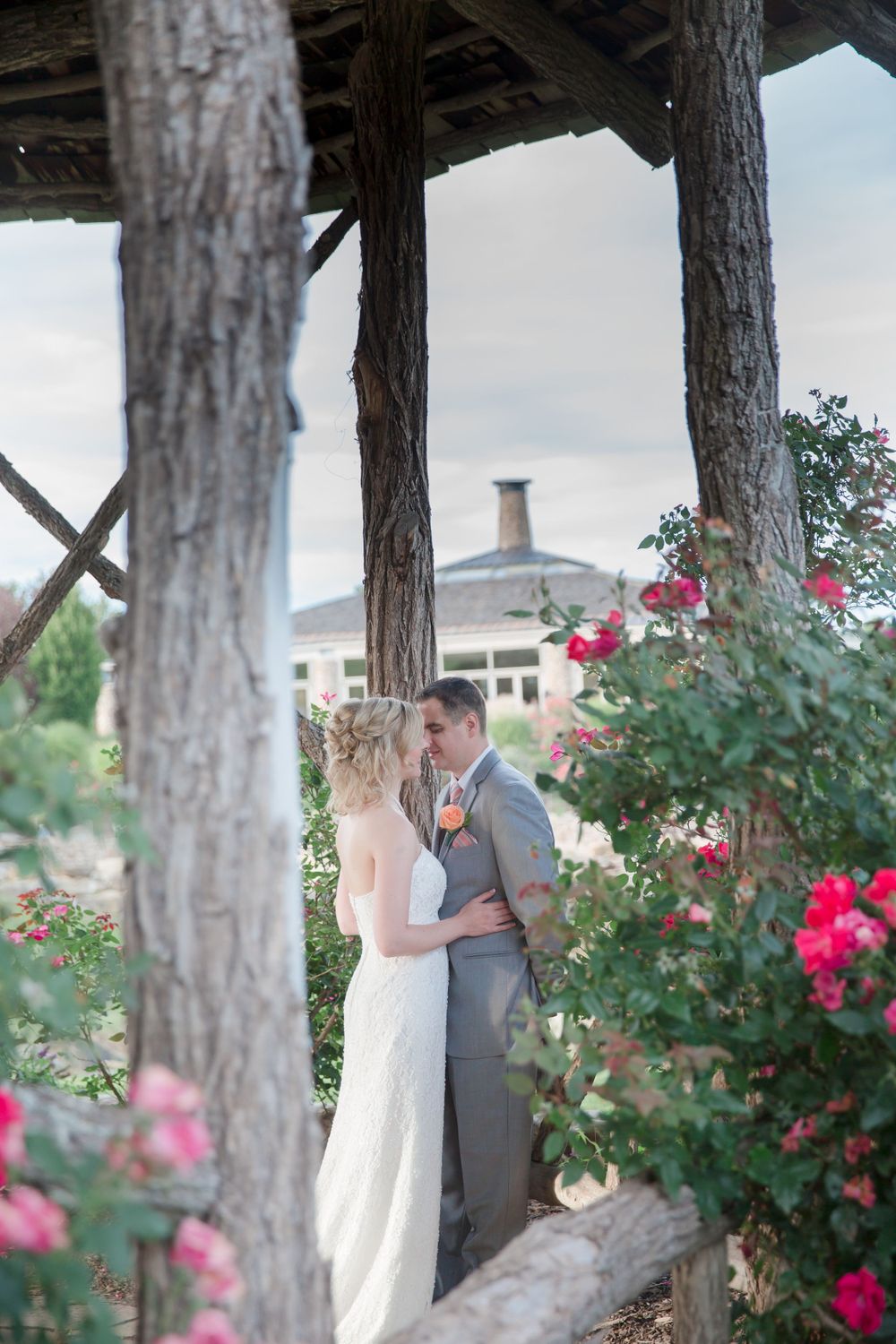 Romantic wedding portrait under wooden pergola with rose garden and building in background.