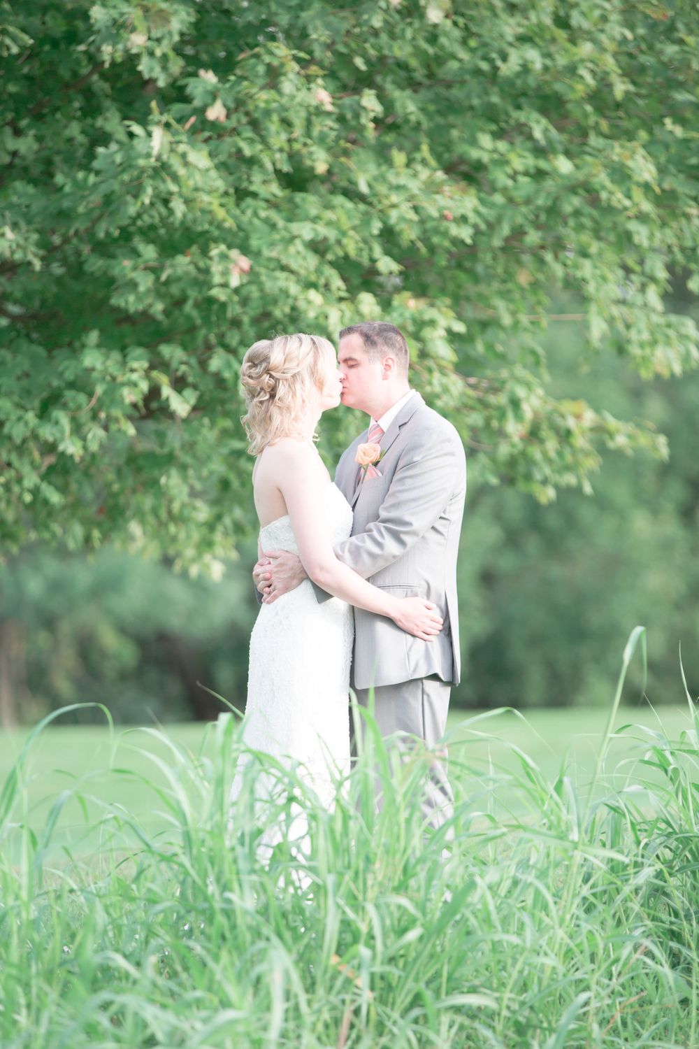 Wedding couple embraces in tall grass with green trees in background.