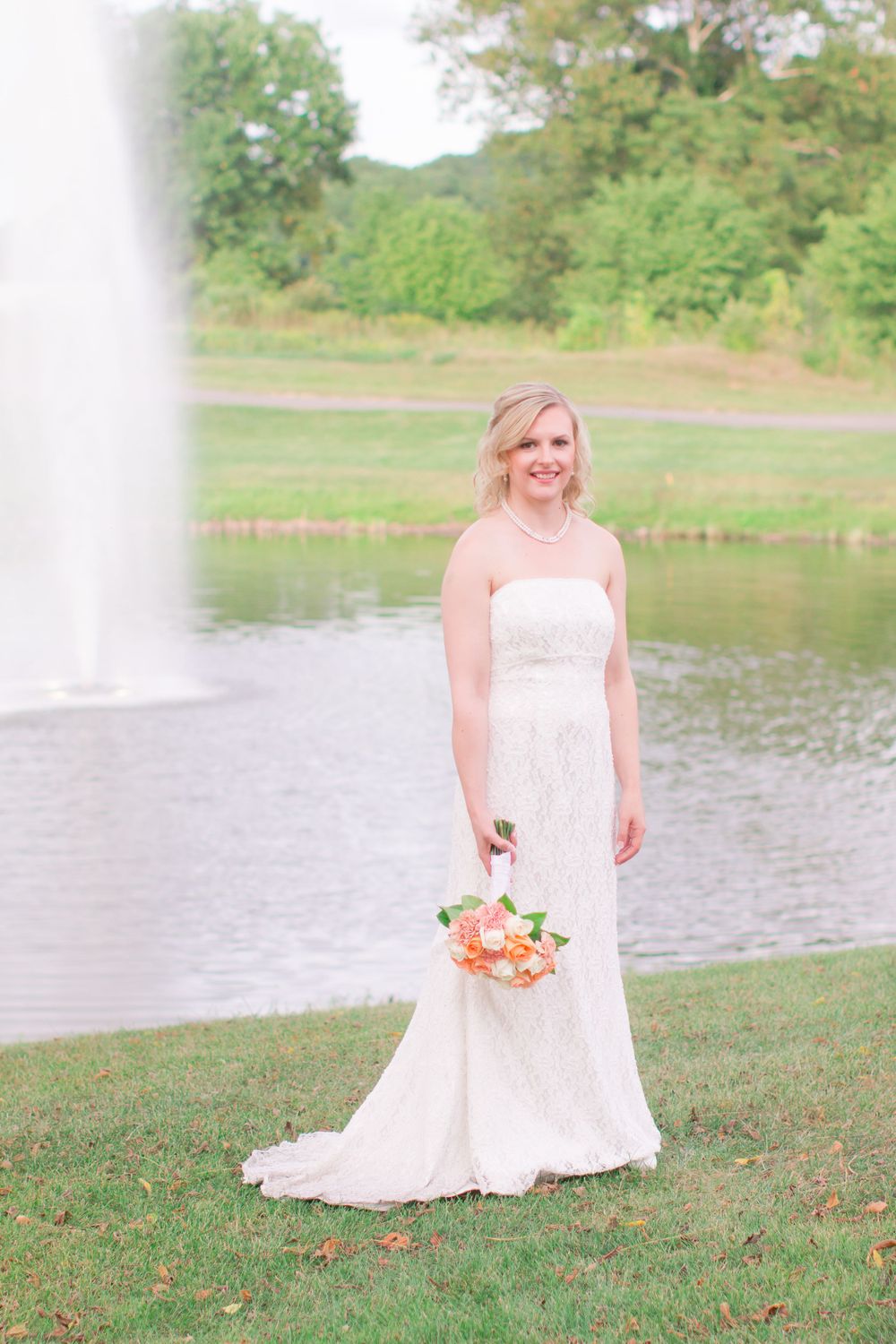 Bride in elegant white dress holds bouquet while standing by fountain and lake.