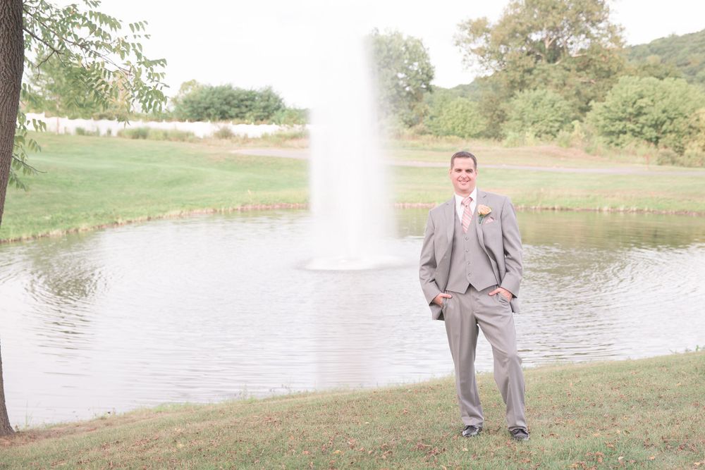 Groom in grey suit stands by fountain and lake on landscaped lawn.