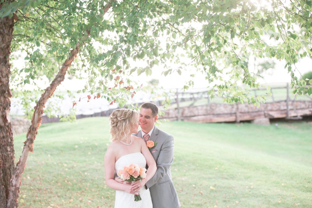 Couple shares romantic moment under tree branches on their wedding day with wooden bridge in background.