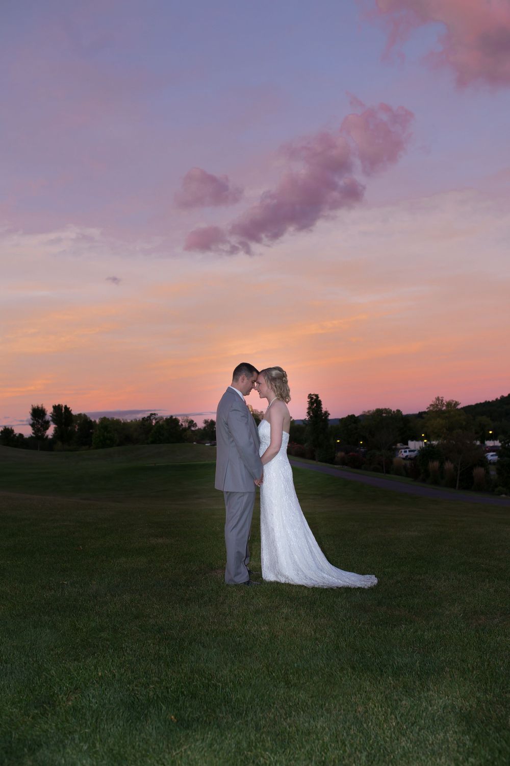 A couple embraces during sunset on a grassy hill with a pink and purple sky.