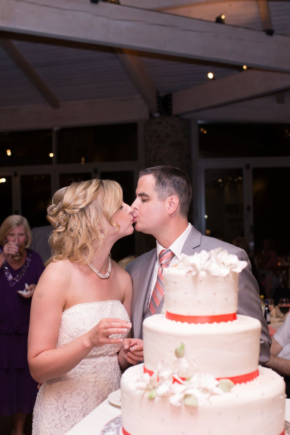 A couple shares a kiss behind their elegant white and red wedding cake at their reception.