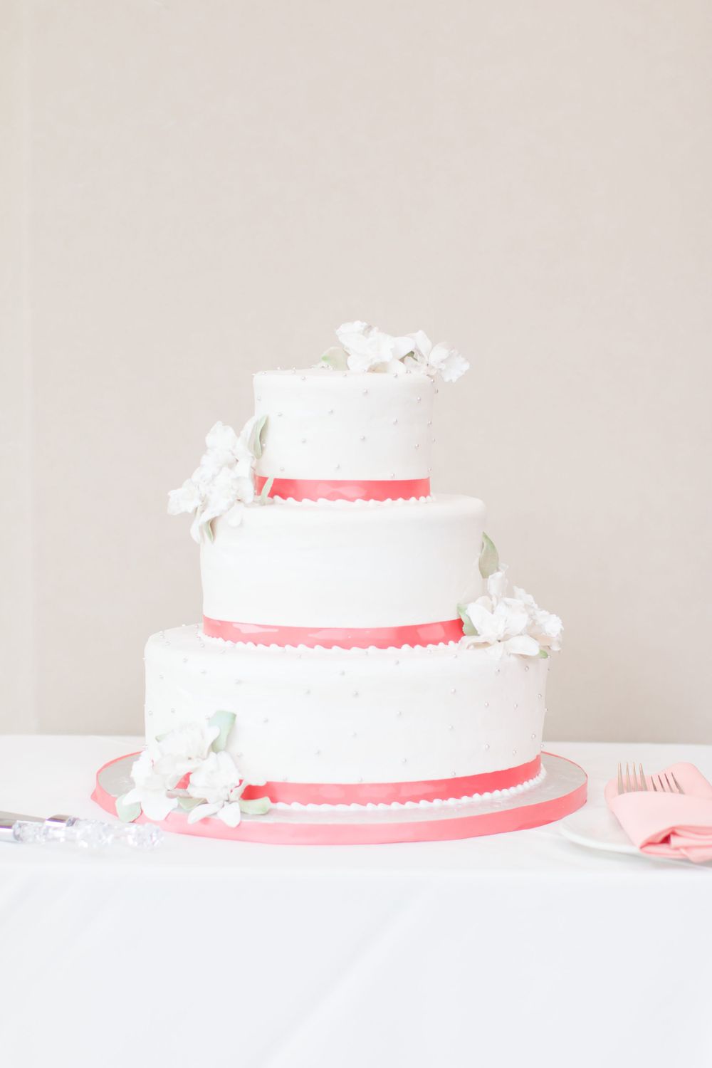 A classic white tiered wedding cake with coral ribbon trim and delicate white flower decorations.