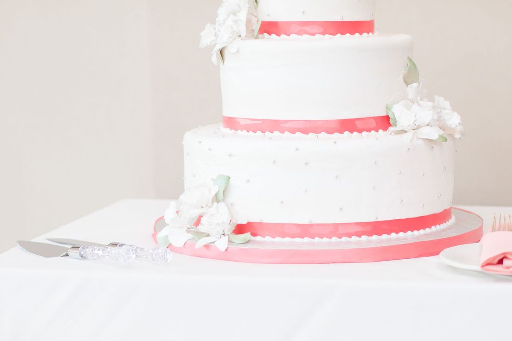 An elegant three-tiered white wedding cake decorated with coral ribbons and white floral accents.