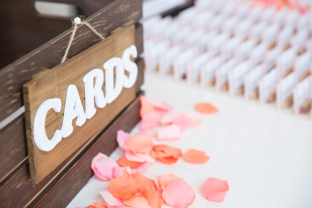 A wooden 'Cards' sign with coral-colored rose petals scattered on a white surface.