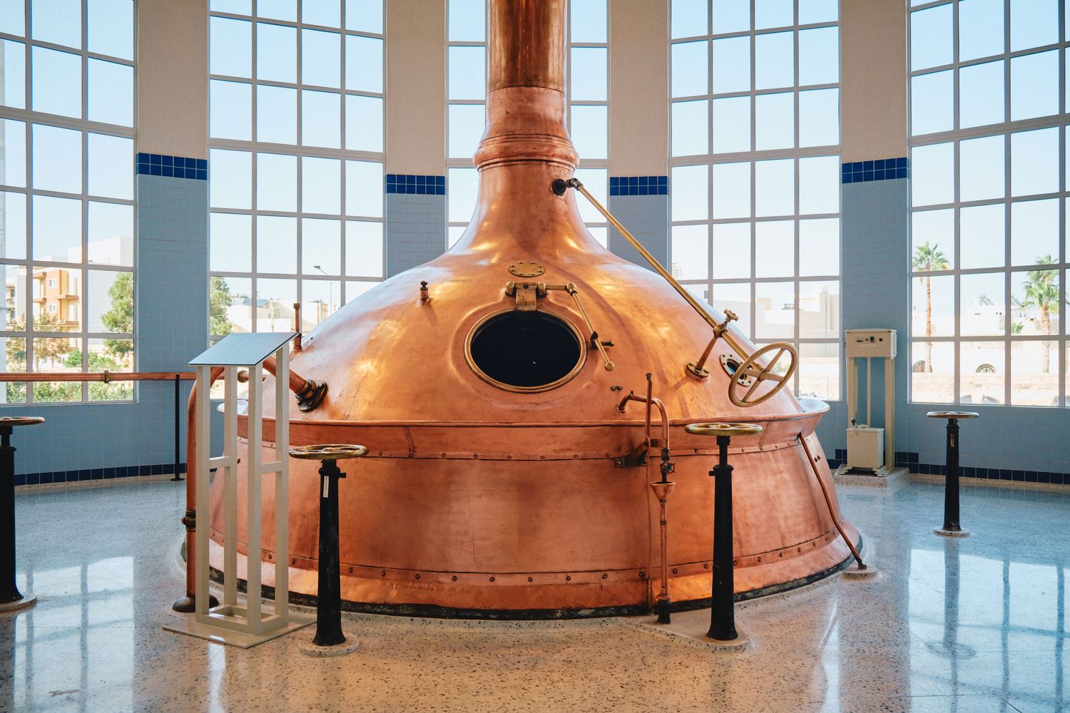 Large copper brewing kettle displayed in modern brewery building with floor-to-ceiling windows.