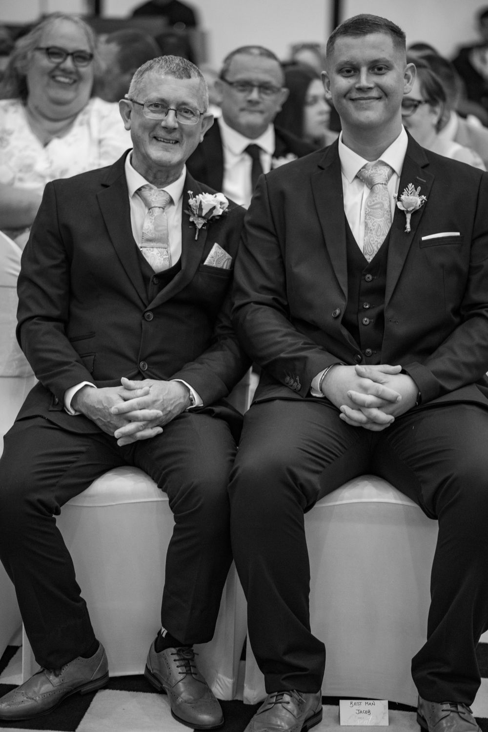 Two well-dressed gentlemen in formal suits sit together smiling at a wedding ceremony.