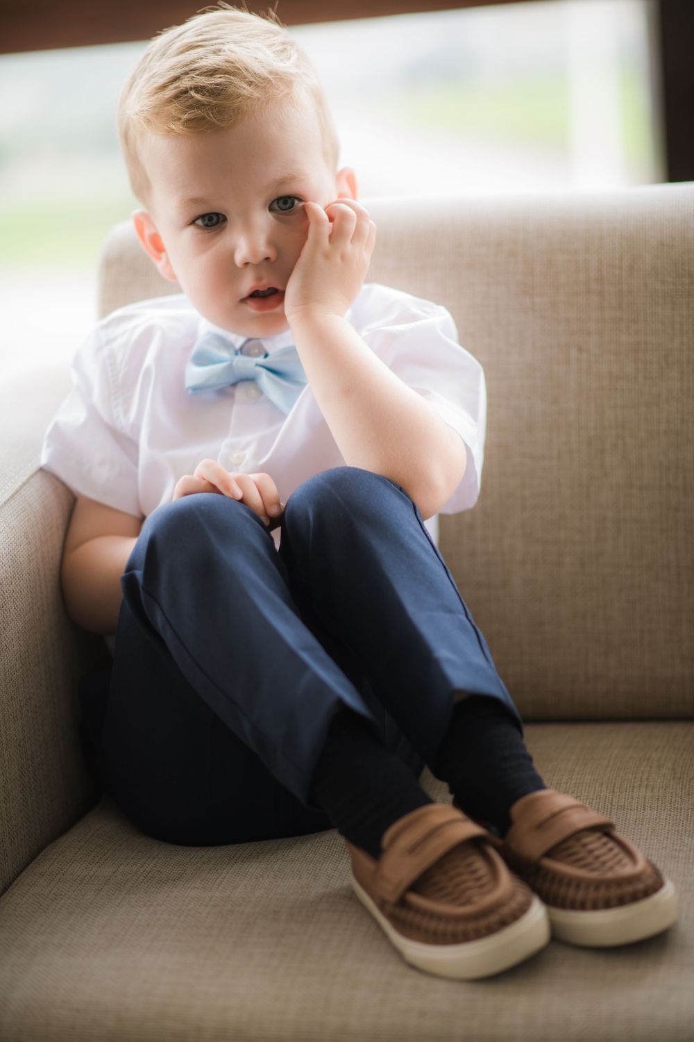 A small child in formal attire with a light blue bowtie sits thoughtfully on a gray couch.