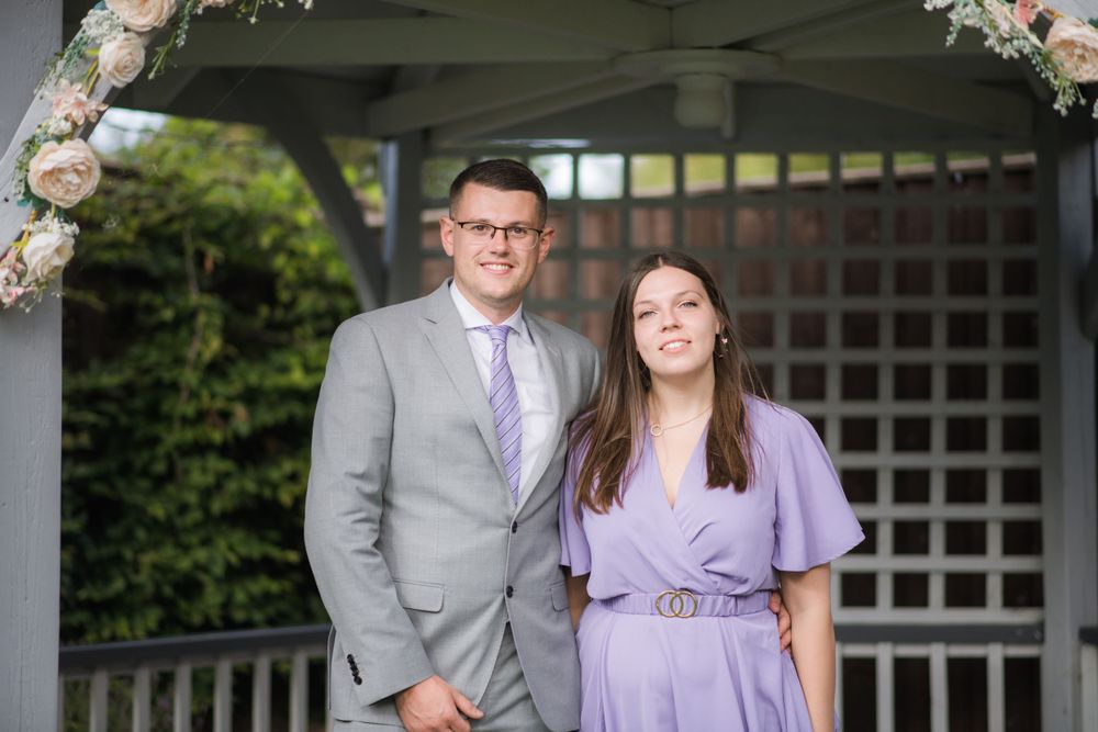 A formal couple poses beneath a decorated gazebo wearing grey and purple attire.