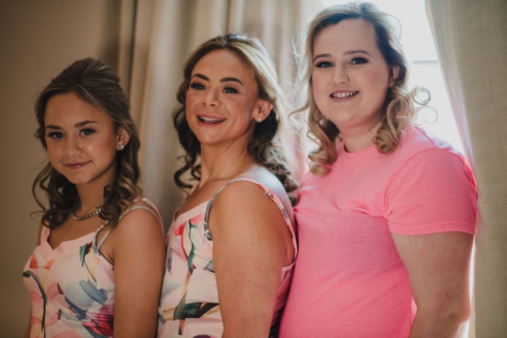Three women in casual and floral dresses pose together by a window.