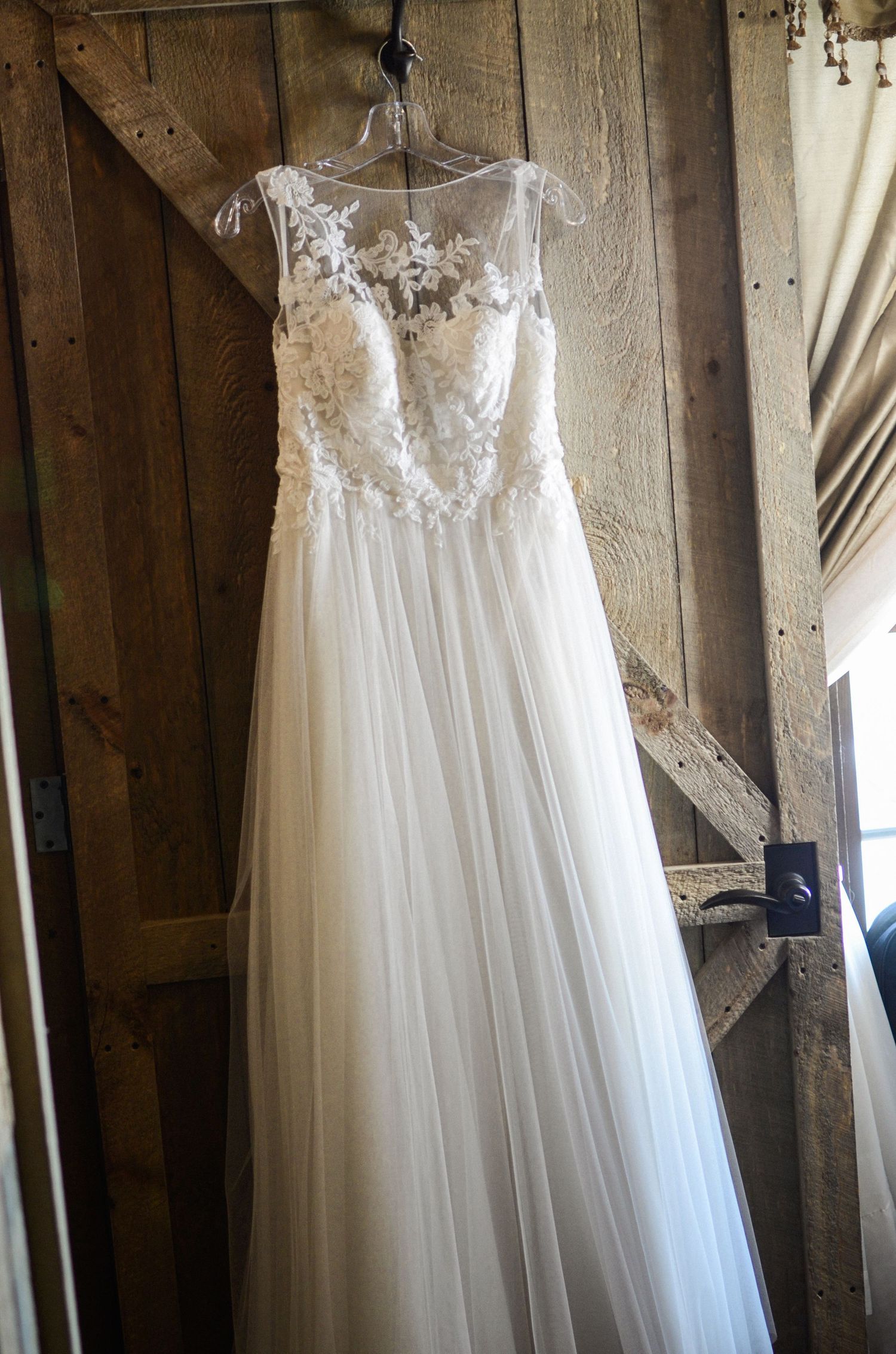 White lace wedding dress hanging against rustic wooden wall.