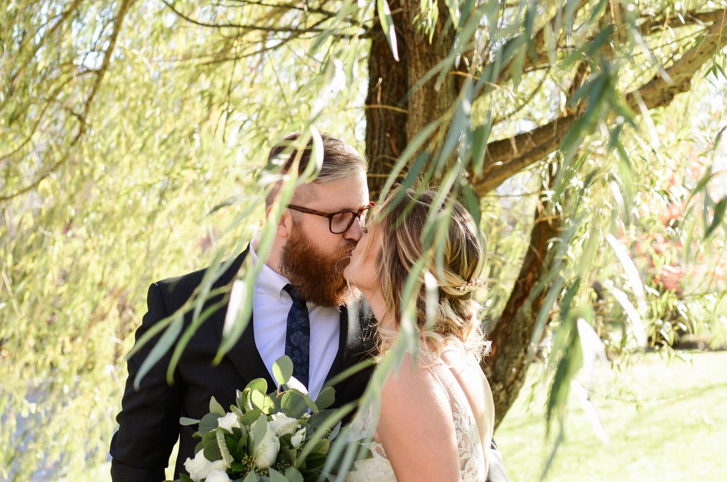 Wedding couple photographed under weeping willow trees in golden sunlight.