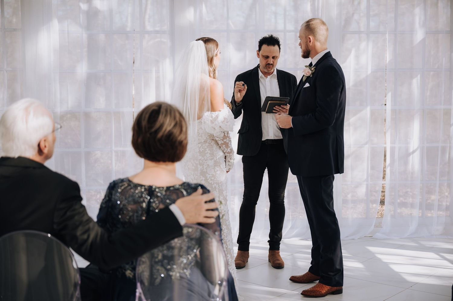 Wedding ceremony in progress within a luminous glass-walled space with seated guests.