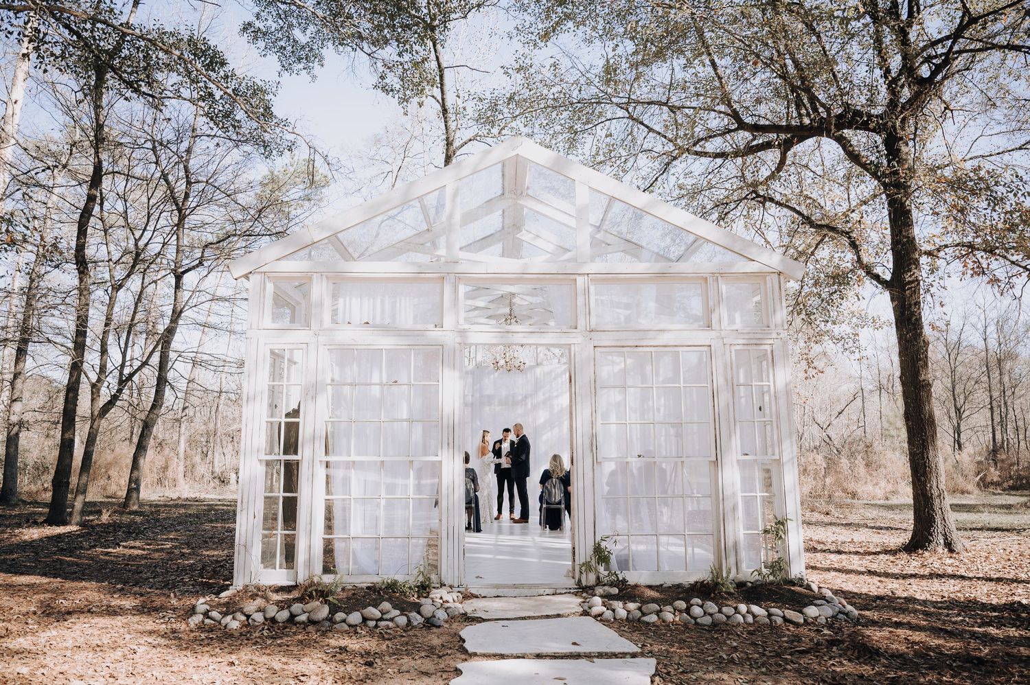 Modern glass chapel surrounded by trees with white interior and stone pathway leading to entrance.