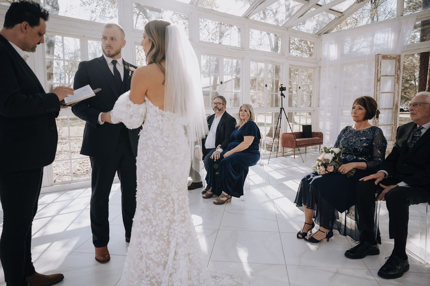 Wedding ceremony with guests seated in a bright glass-enclosed venue with white decor.