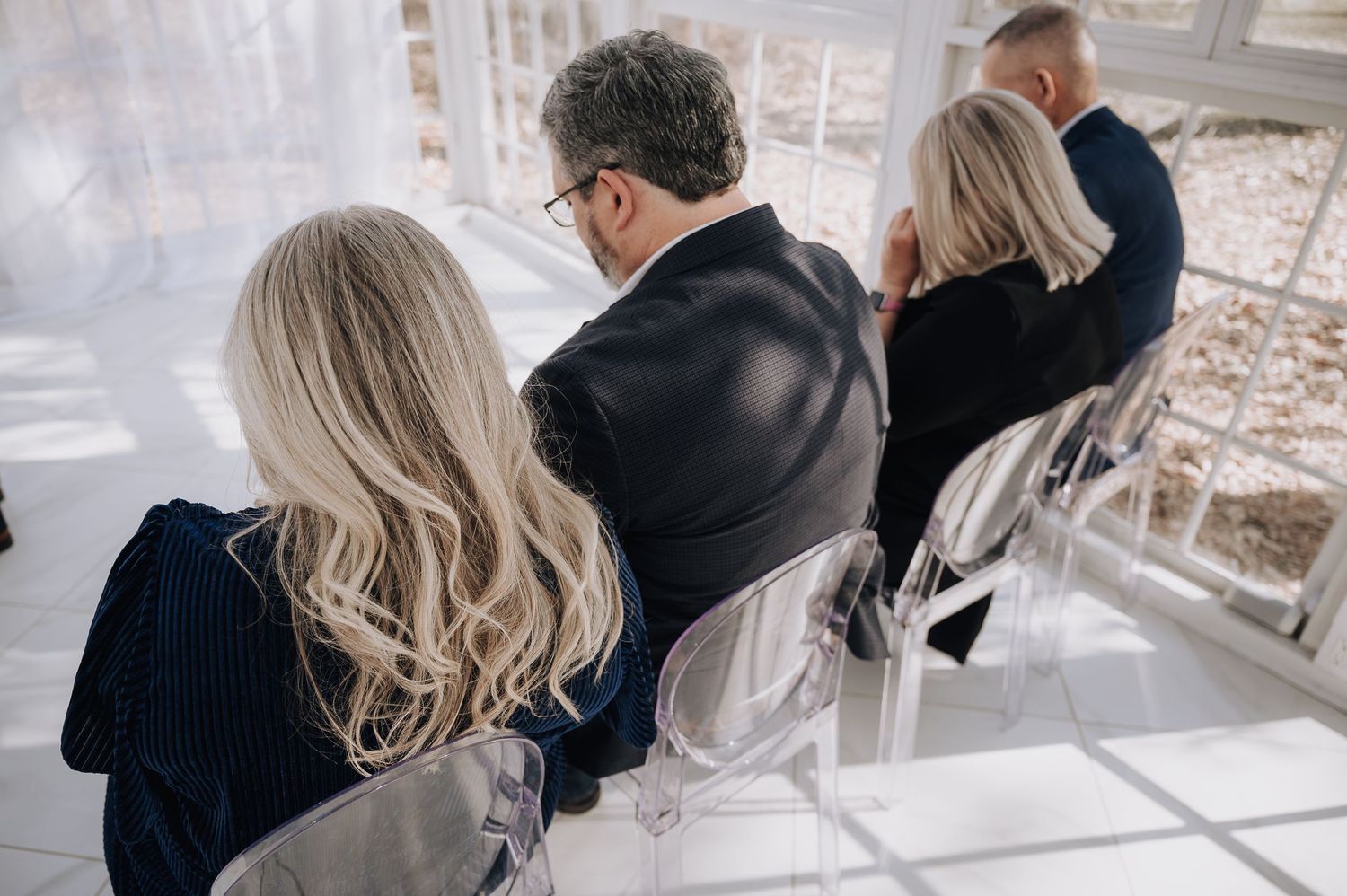 Wedding guests seated in white chairs inside a bright glass-enclosed ceremony space.