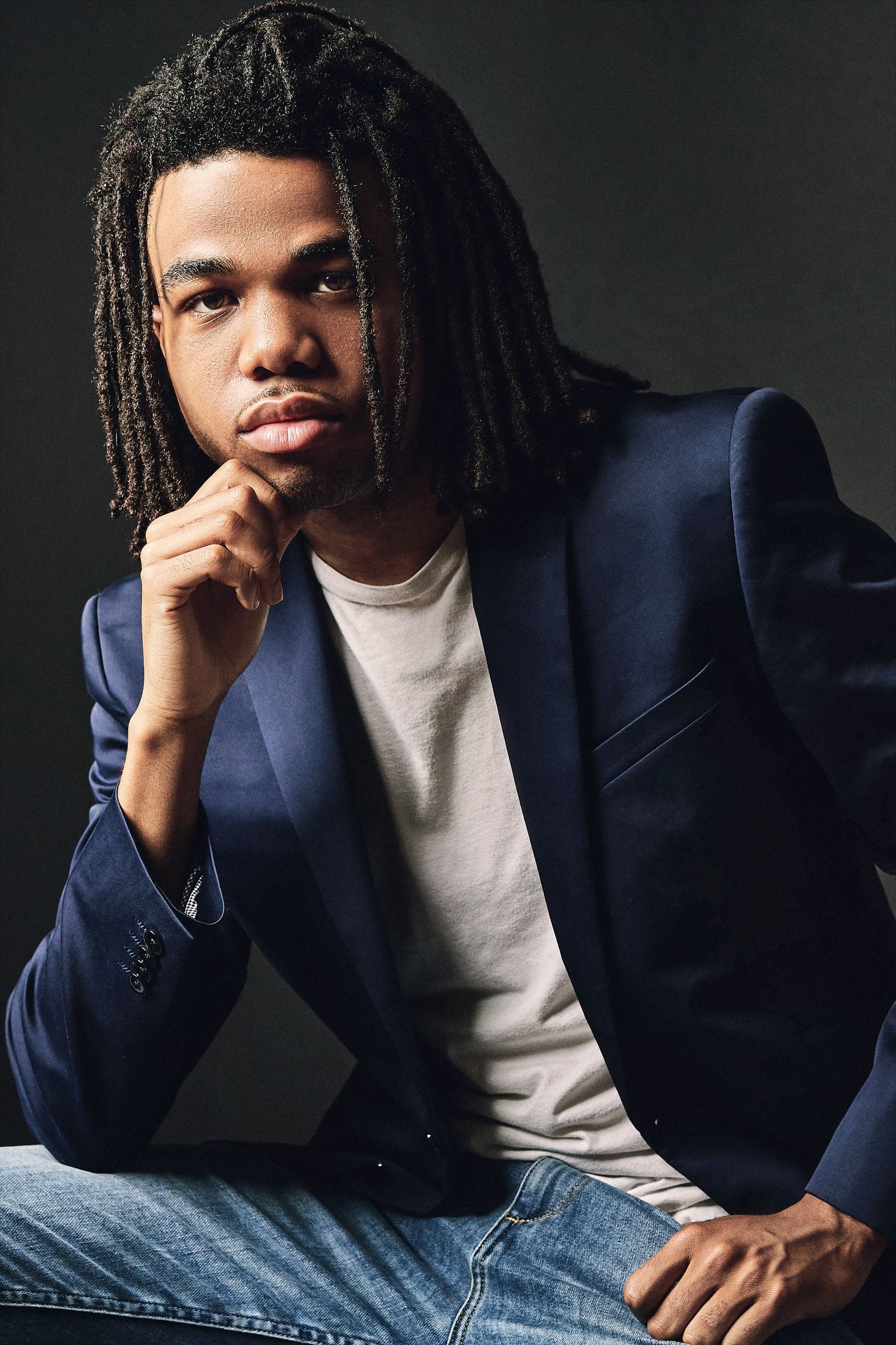A young man with dreadlocks in a navy blazer and jeans poses thoughtfully against a dark background.