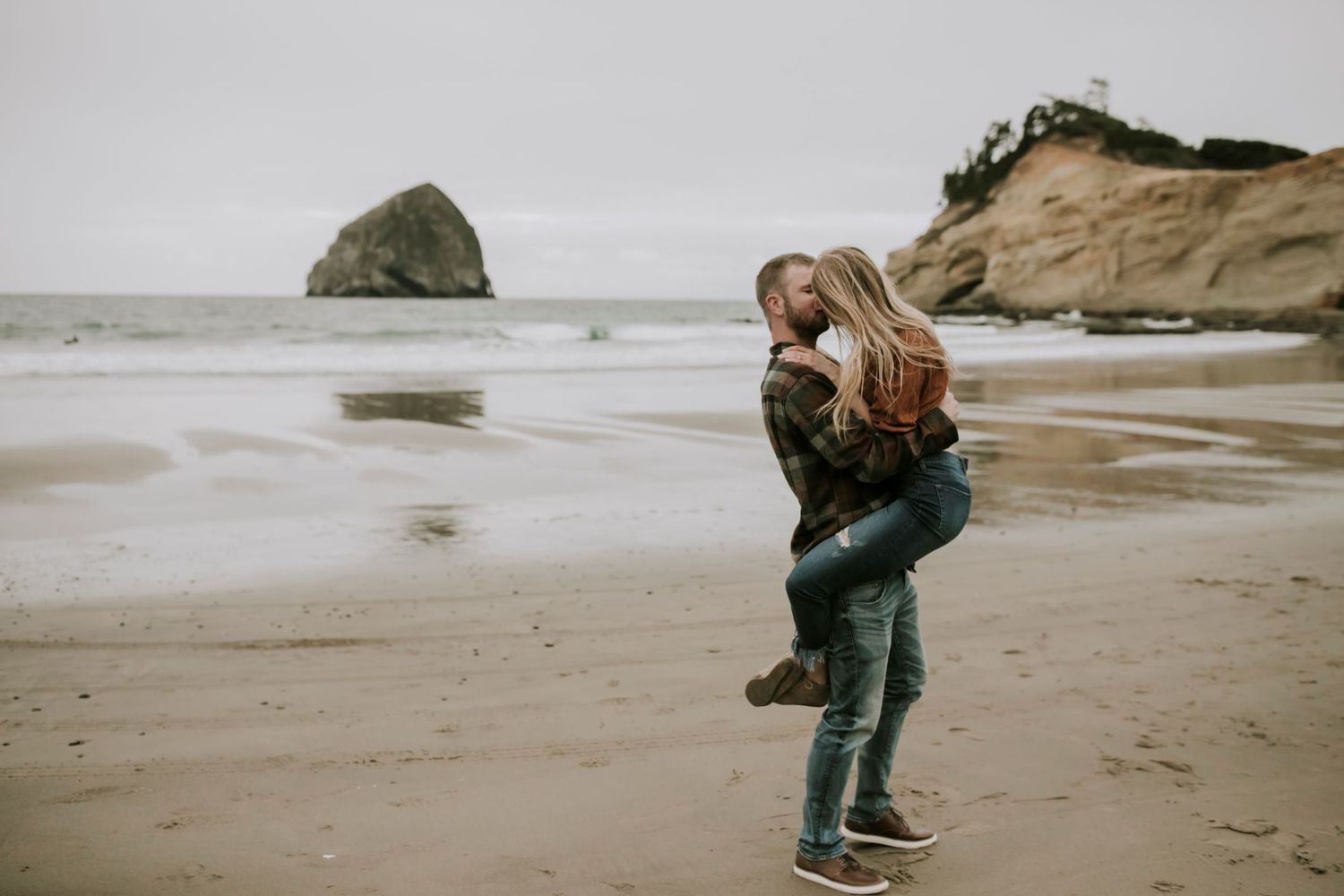 Boy holding girl while kissing on the beach with ocean behind them in Oregon