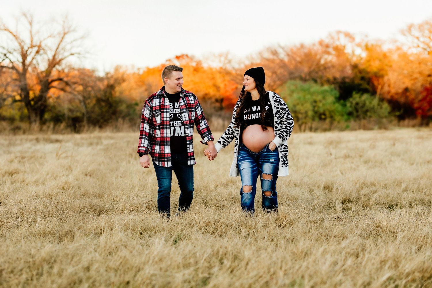 Expectant couple in casual plaid shirts walking through autumn field at sunset.