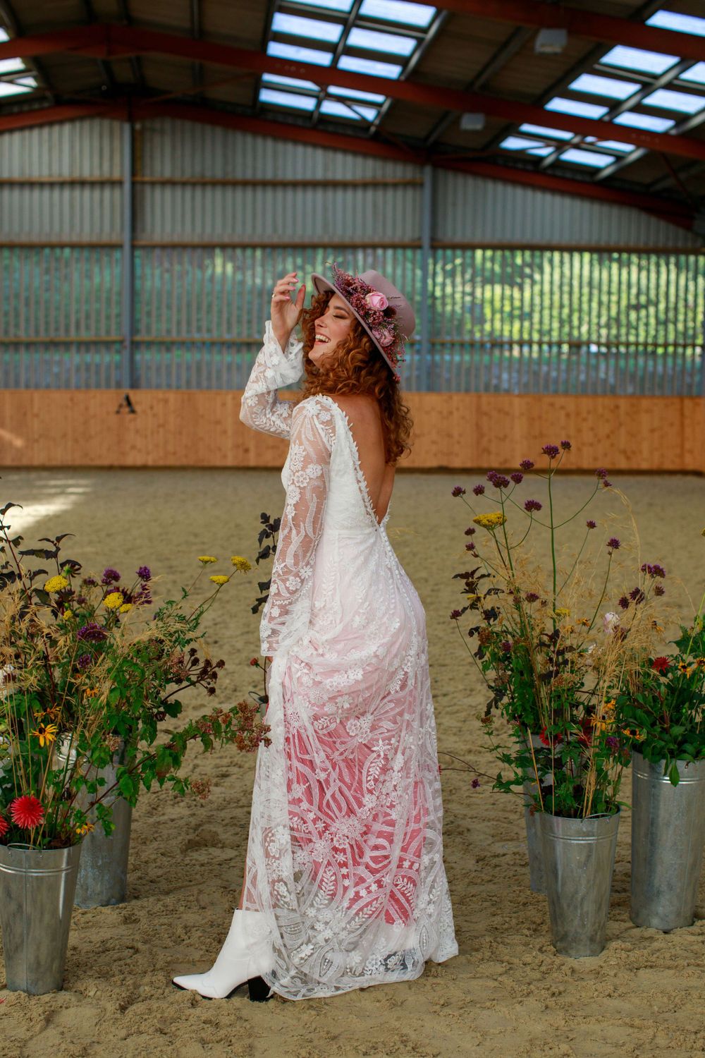 April stood in a horse arena with flowers next to her wearing a wedding dress and cowboy hat and boots