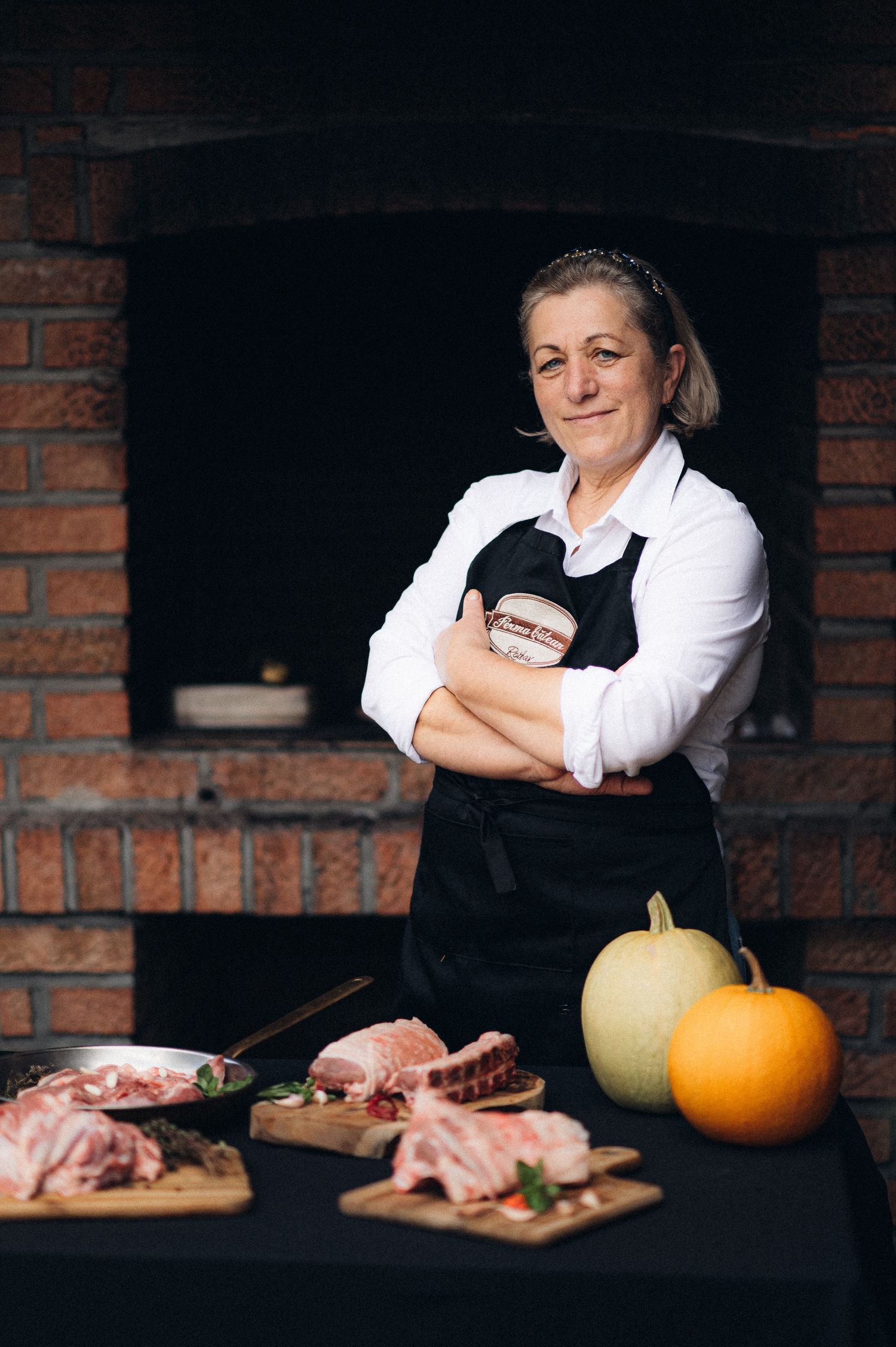 Chef in black apron and white shirt stands near brick oven with pumpkins and prepared meat.