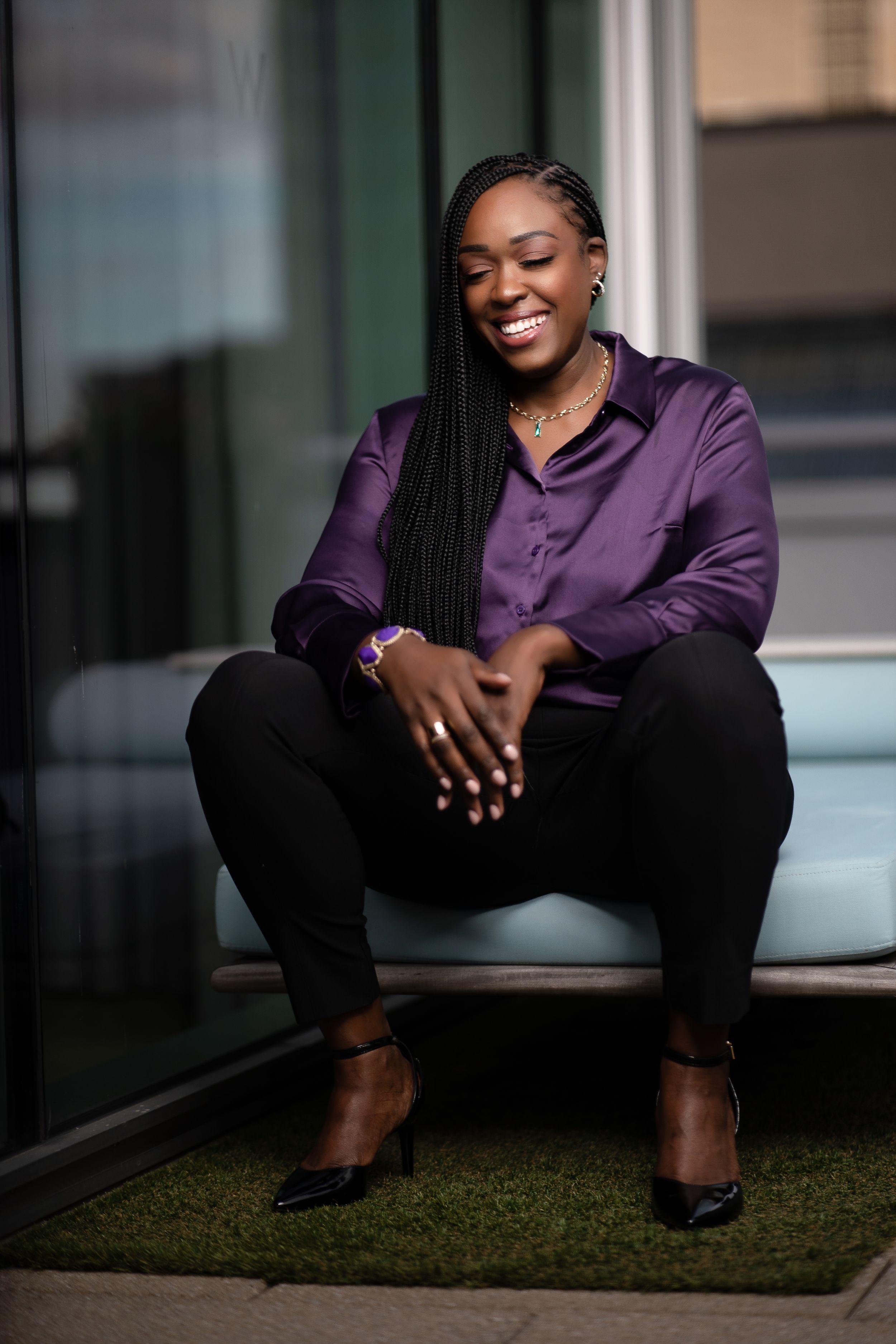 Smiling woman in purple satin shirt sitting relaxed outdoors with braided hair and black pants.