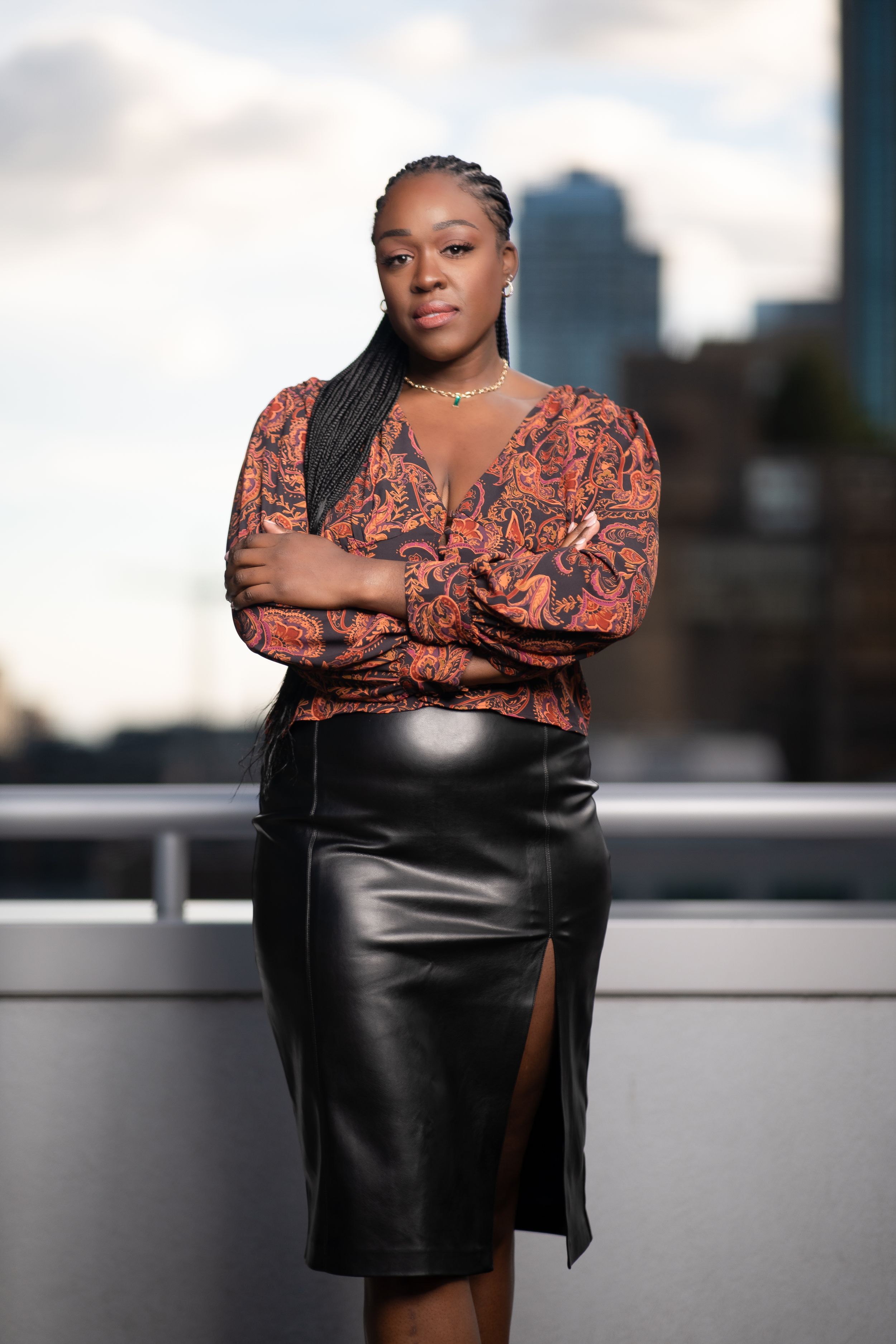 Woman in floral blouse and black leather skirt posing confidently on a rooftop with city skyline.