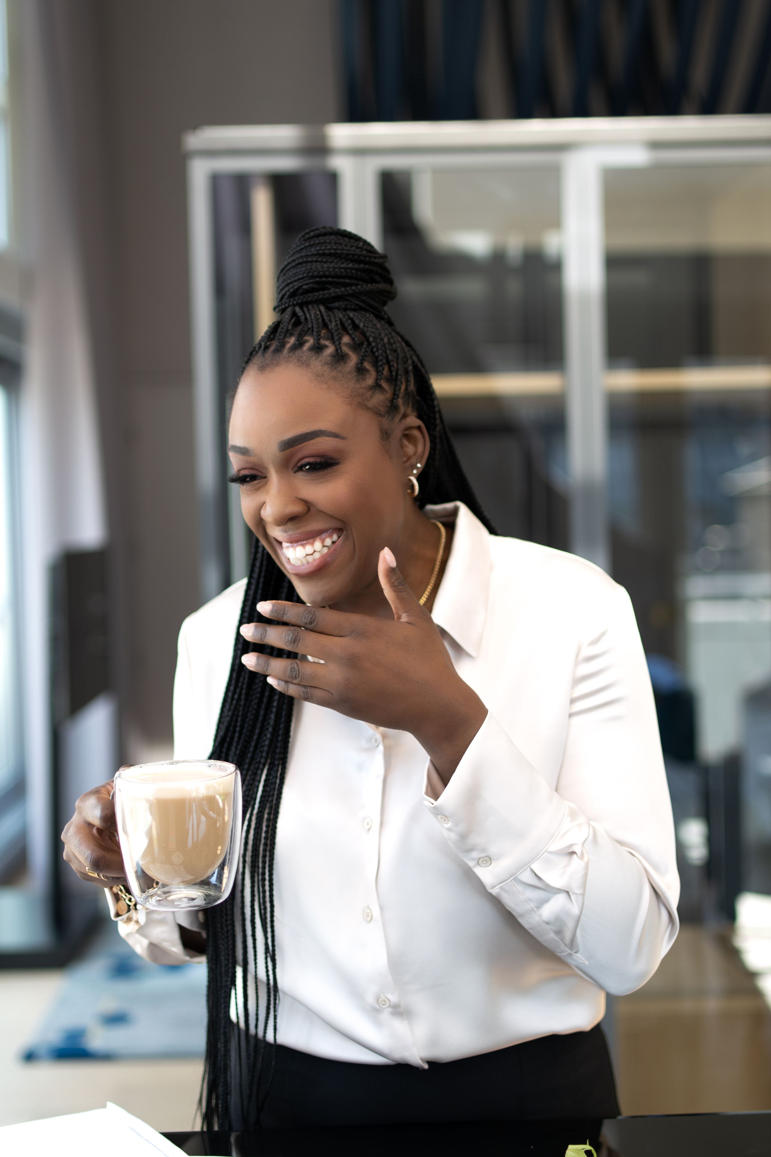 Laughing woman in white blazer holding a glass coffee drink indoors with hand on chest.