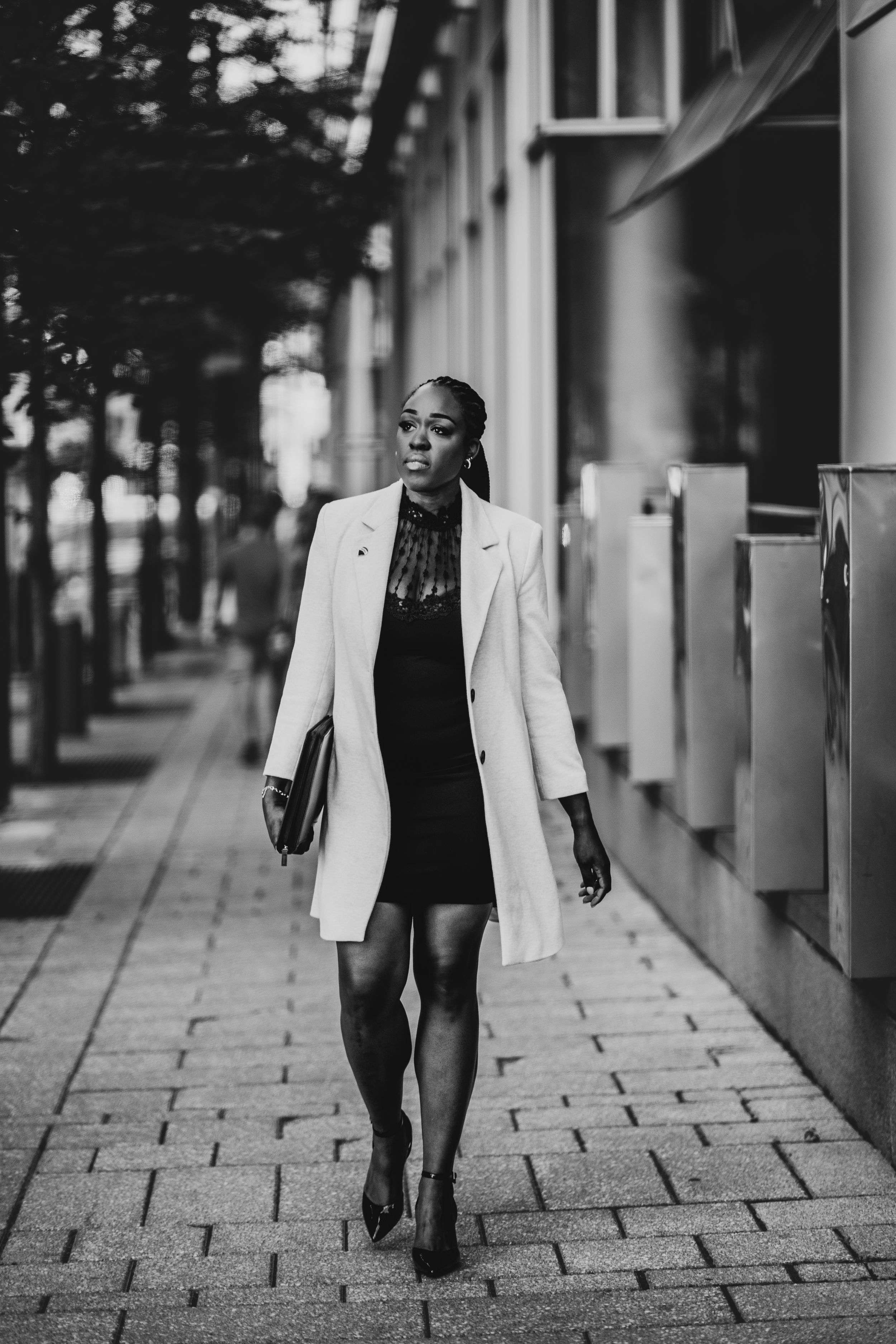 Black and white photo of a woman in white coat walking confidently along an urban sidewalk.