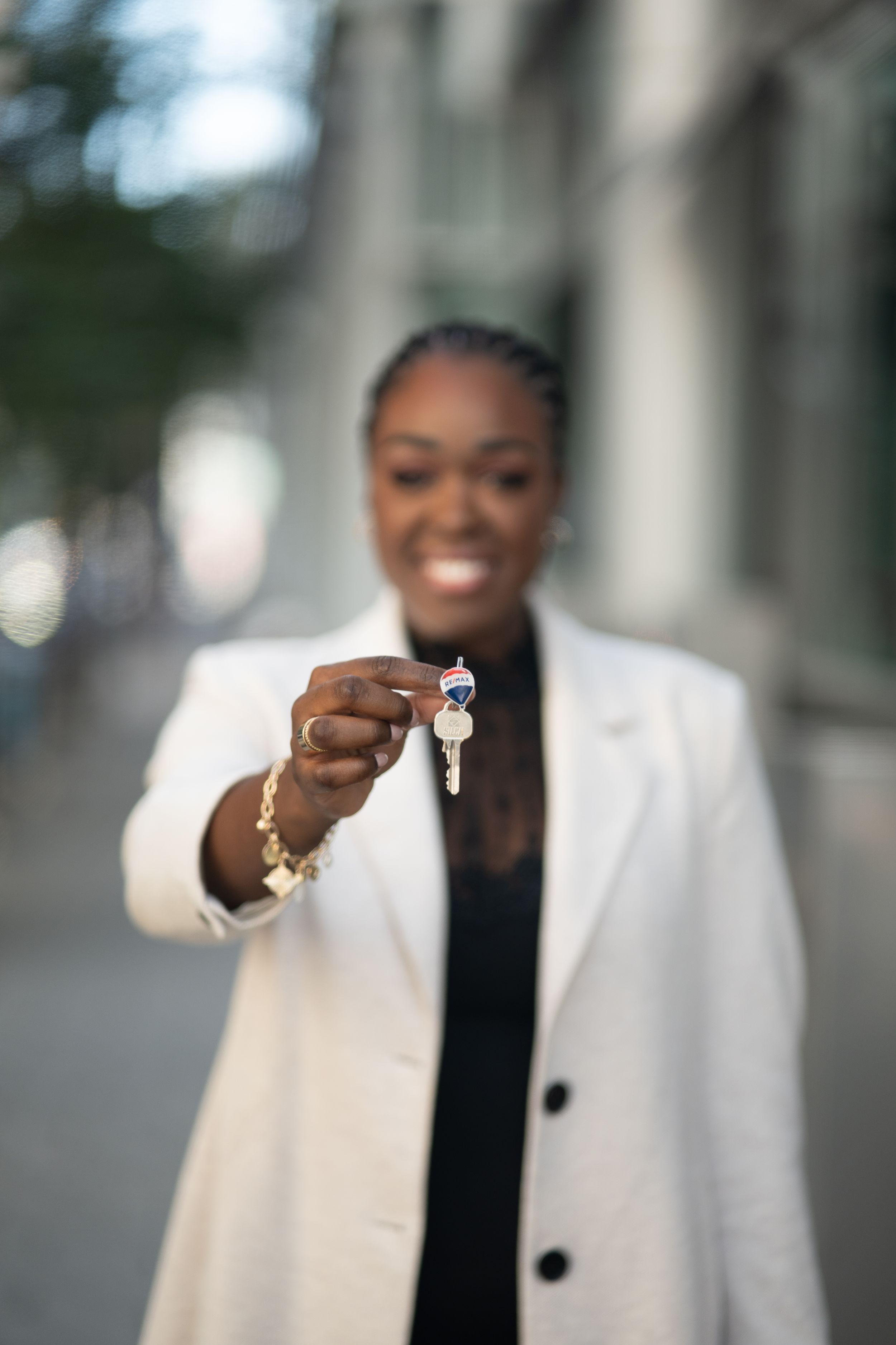 Woman in white blazer holding RE/MAX real estate keys toward camera on a city street.