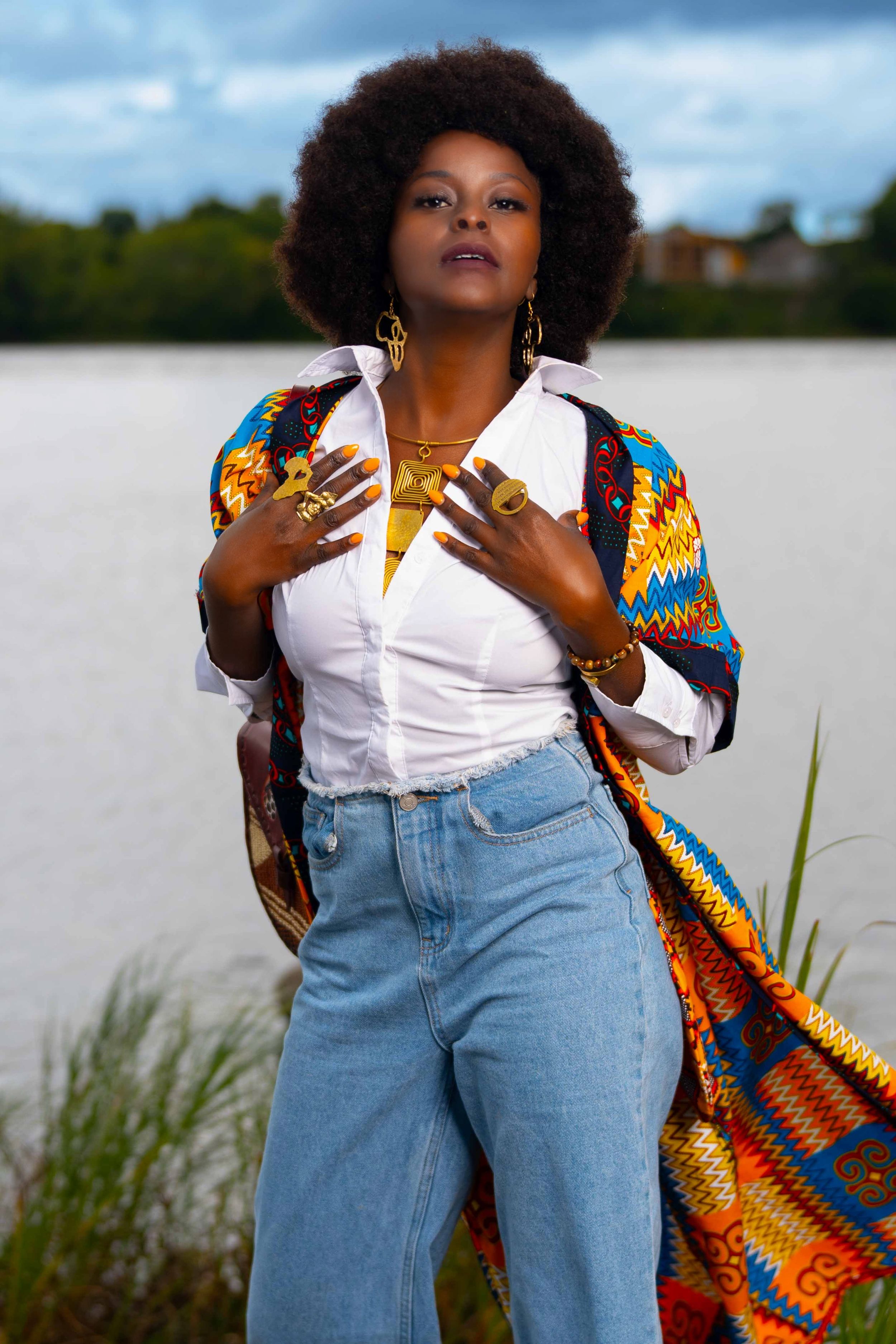Woman with natural afro wearing colorful kente kimono, white shirt, and jeans near a lake.