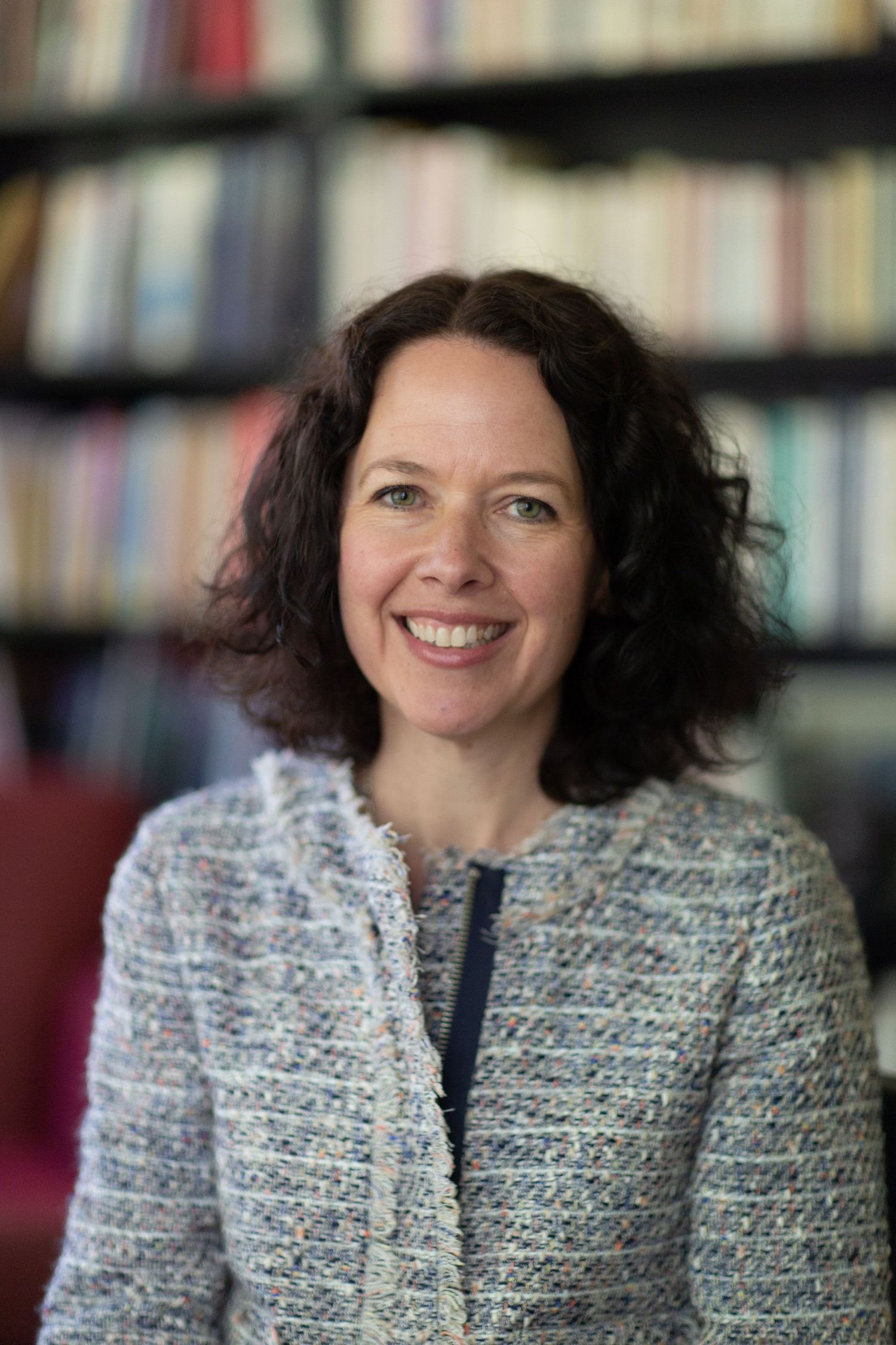 A person wearing a textured grey tweed jacket poses in front of a bookshelf.