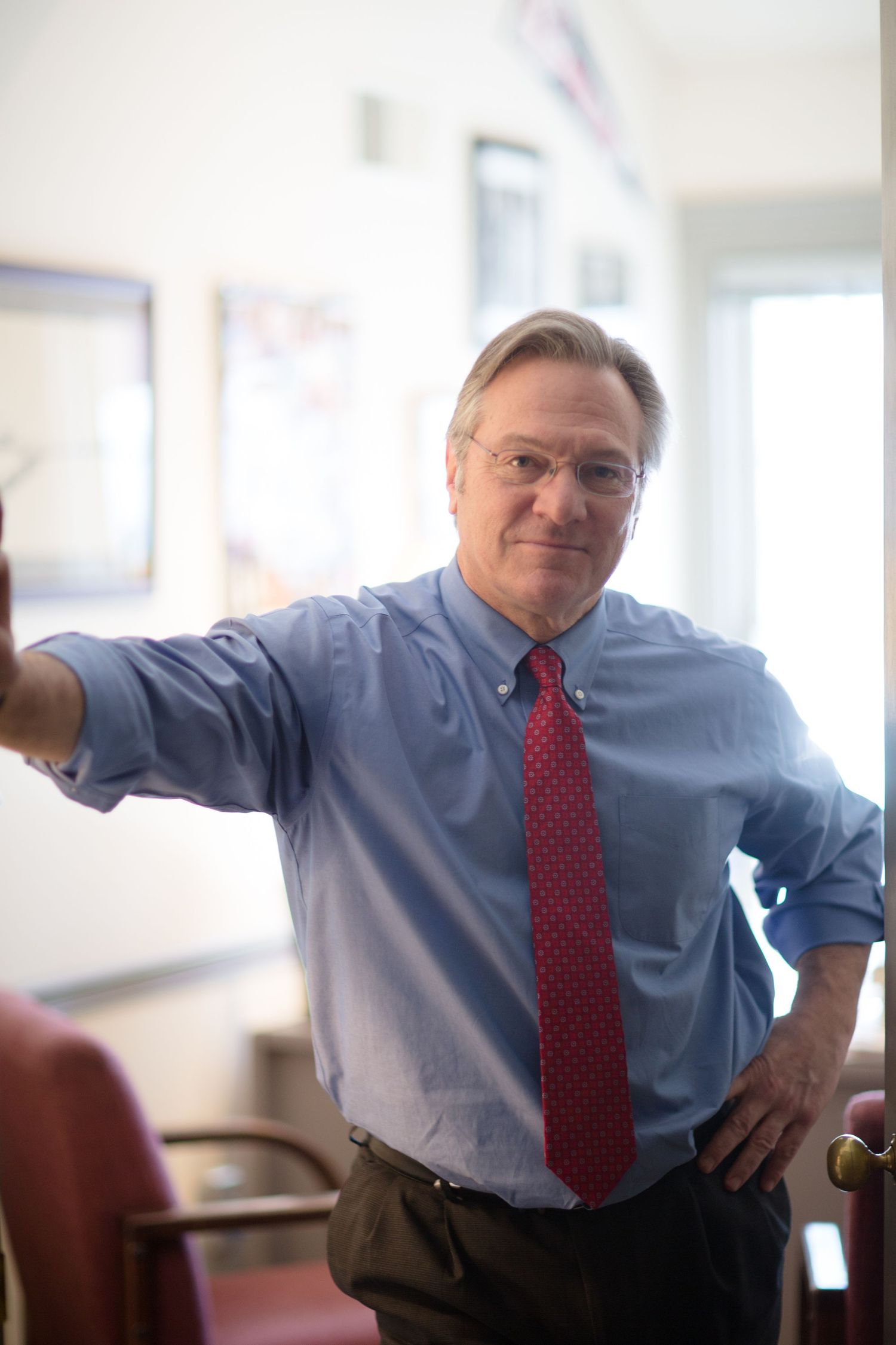A person in a blue dress shirt and red tie leans against a bright office space.